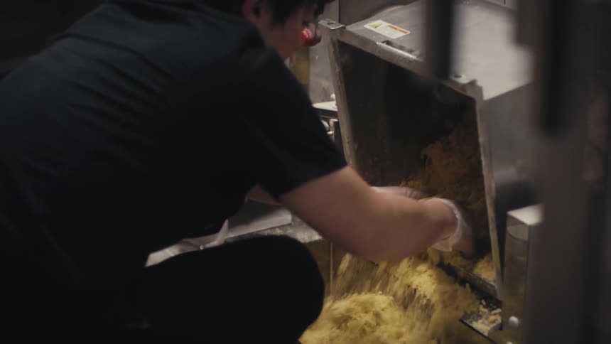 Closeup of a woman with gloved hands scooping and breaking a handful of dough in a metal container