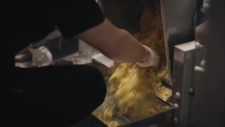 Closeup of a man with gloved hands scooping and breaking a handful of dough in a metal container