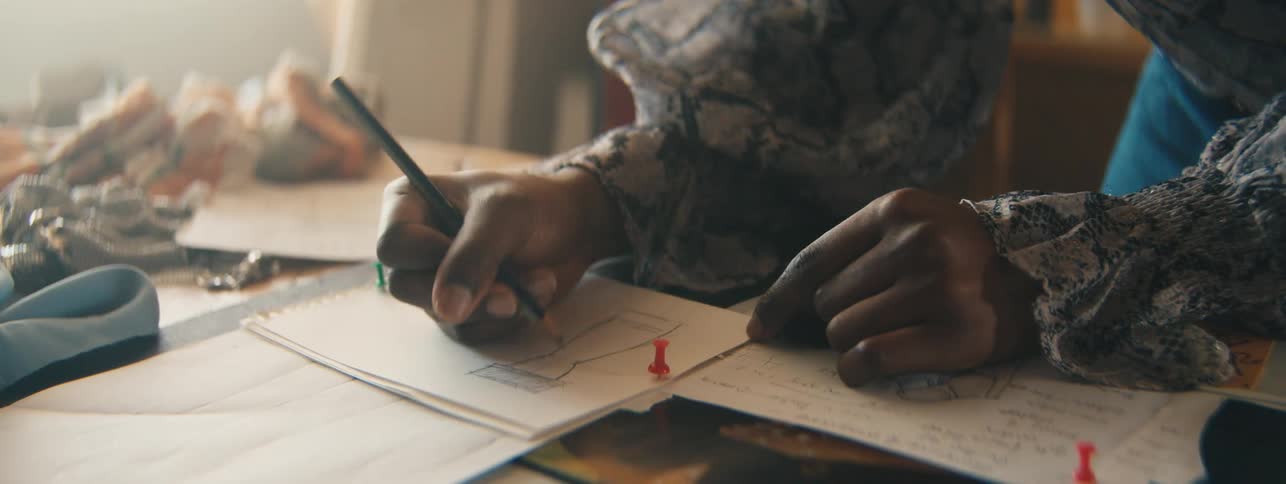 The human hands of a person holding a dark pencil and drawing on a white paper and the persons sleeves are wearing a long dress