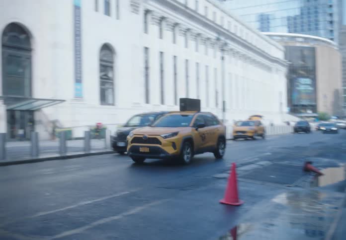 A taxi driving down a midtown street with cars parked on the sides and Madison Square Garden in the background
