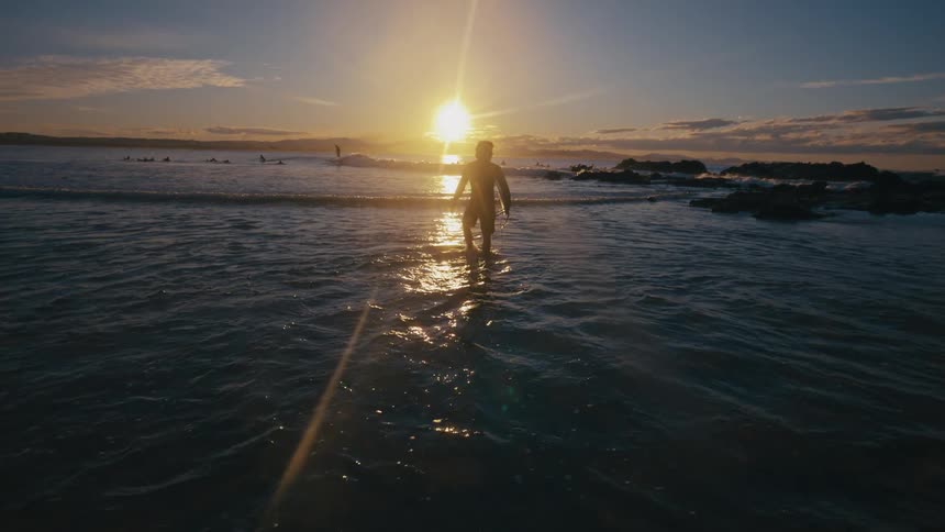 A surfer carrying a surfboard walks towards the ocean at sunset