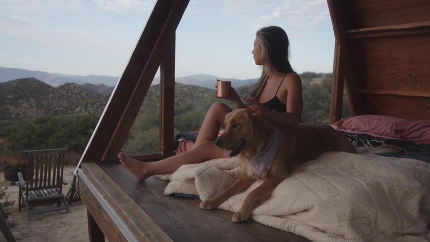 A woman sits on a bed in a small wooden structure in the desert drinking from a cup while her dog lies beside her