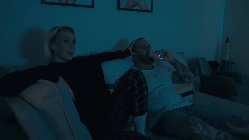 A man and a woman are sitting on a couch looking forward where there is a screen that is illuminating the dark room they are in and the man is drinking something from a can