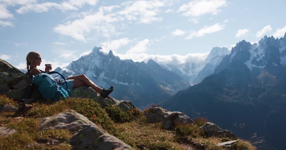 A woman sits on a rocky mountainside sipping a drink and taking a break from hiking