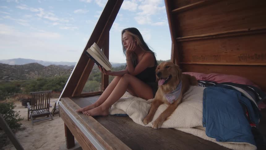 A woman sits on a bed in a small wooden structure in the desert reading a book while her dog lies beside her