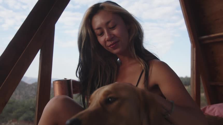 Woman sitting in a cabin drinking a cup of coffee and petting a golden retriever next to her