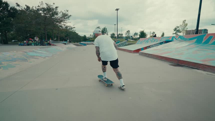A man wearing a white shirt and black shorts rides a skateboard across a concrete skatepark