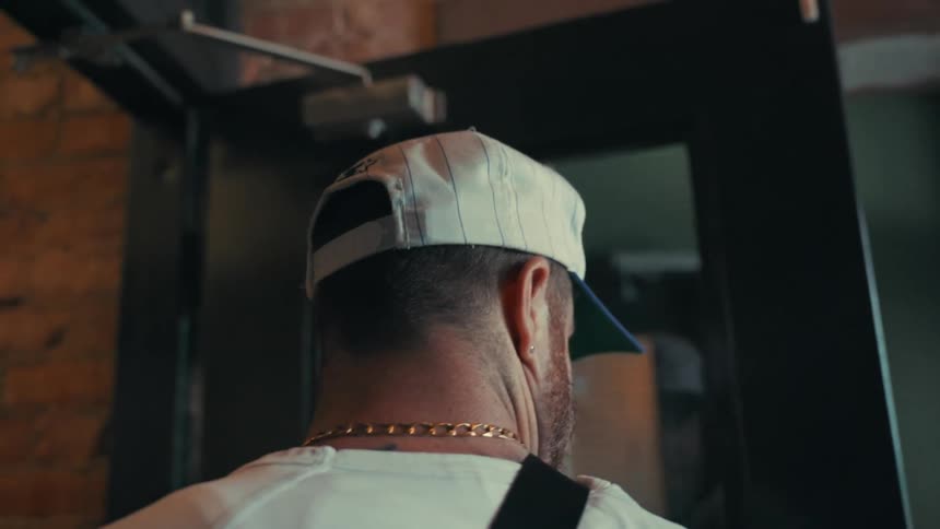 A closeup of a mans back wearing a baseball cap and a gold chain as he enters a building