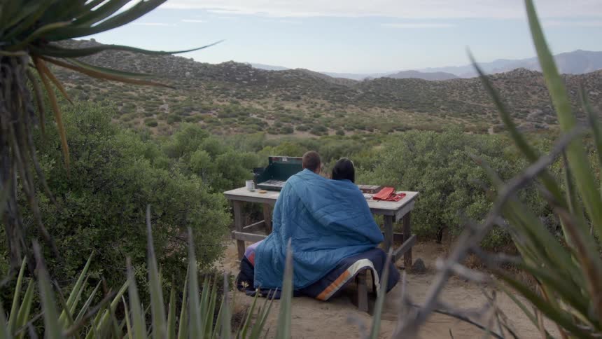 Back view of a man and a woman sitting on a blue blanket amidst hills with undergrowth
