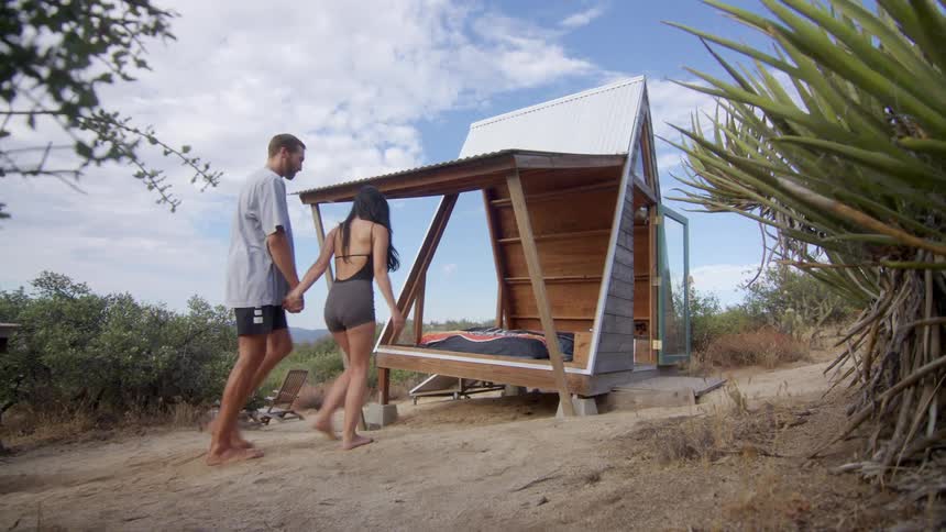 Man and woman walk hand in hand towards a micro cabin with an open side on a cloudy summer day