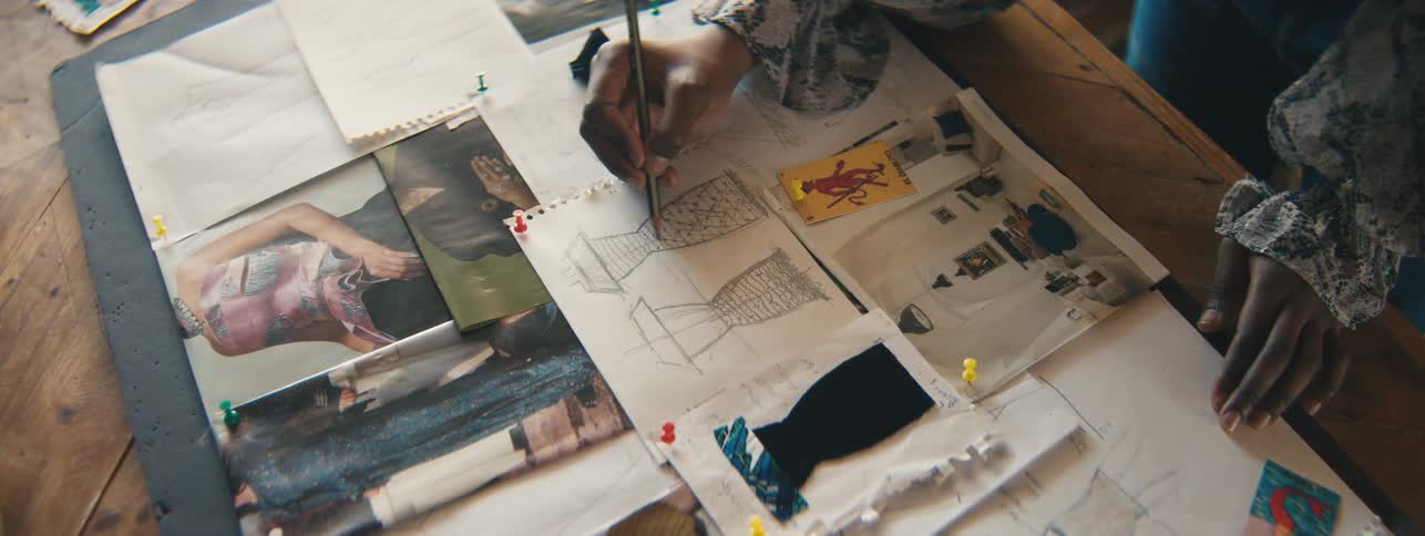 A persons hands using a pencil to draw a dress on white paper on a table full of pictures of dresses