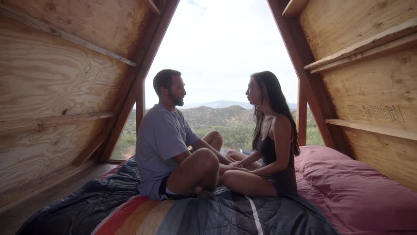 Couple sitting facing each other on the bed in a cabin with mountains in the background