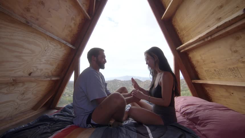 Man and woman sitting in a wooden cabin holding hands against a background of mountains