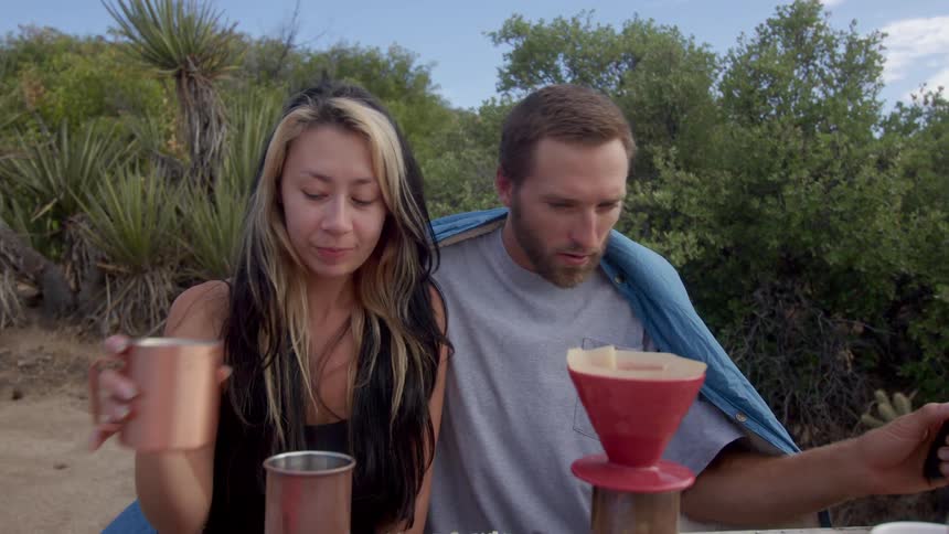 Woman kissing a man who is preparing coffee while they are on a hill surrounded by greenery