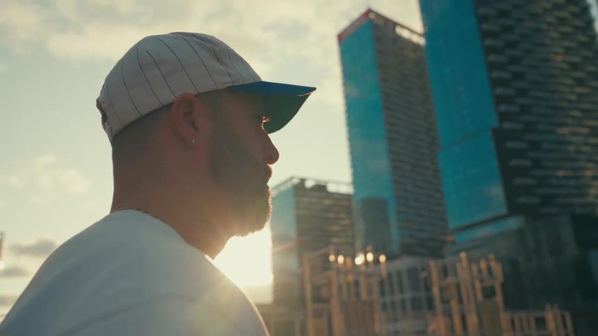 The face of a young bearded man with a white cap walking forward and there are some large office buildings