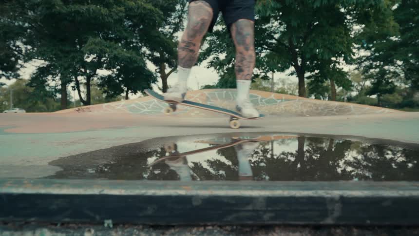 A skateboarder with heavily tattooed legs rolls past a puddle in a skatepark