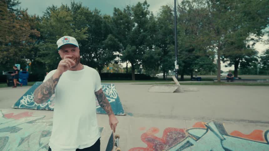 A man in a white shirt and cap is walking forward while smiling and he is holding a skateboard and he is on a skate park with various graffiti