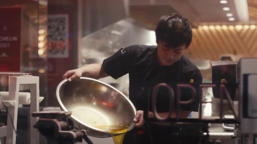 View from outside a restaurant of a chef holding a metal bowl and pouring oil