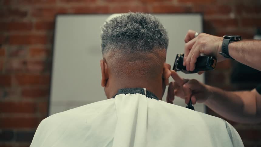 The back of a mans neck having his hair cut by a man standing to the right using a hair clipper