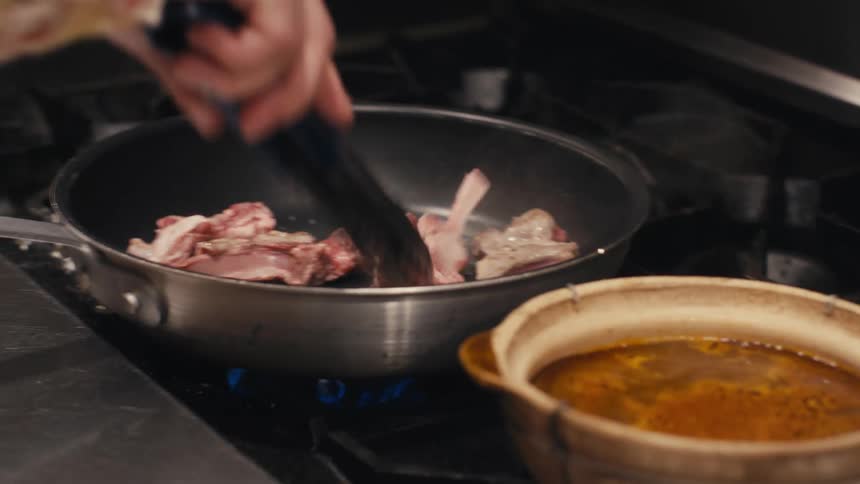 A hand with tongs cooking meat in a pan next to a bowl of hot soup