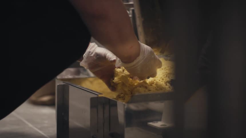 Closeup of gloved hands scooping and breaking a handful of dough in a metal container