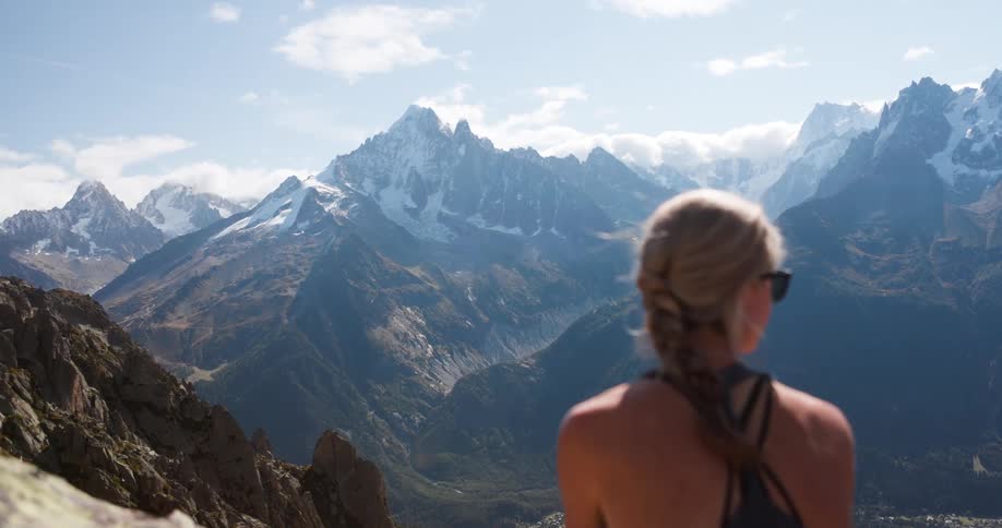 A blonde woman wearing sunglasses sits at the top of a mountain and takes in the view of a mountain range and valley