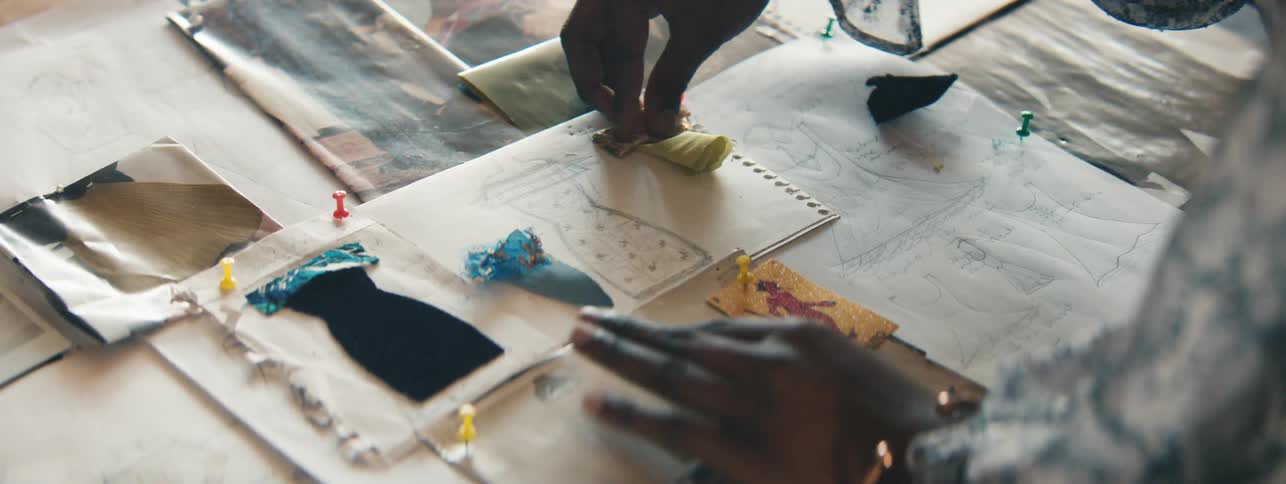 Hands of a female fashion designer placing clothing fabrics in the shape of dresses on drawings that are on the table