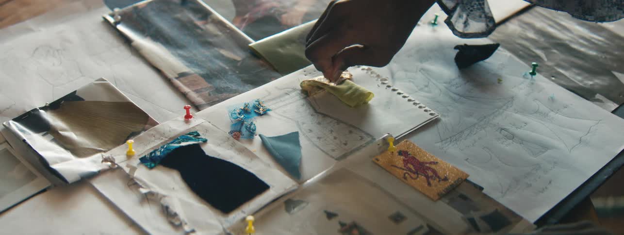 A persons hand using a pin to pin a piece of fabric on top of some papers with design drawings