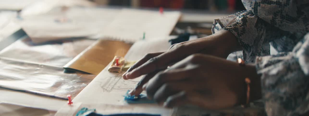 Hands of a female fashion designer placing fabric dresses on top of drawings on white papers on the table