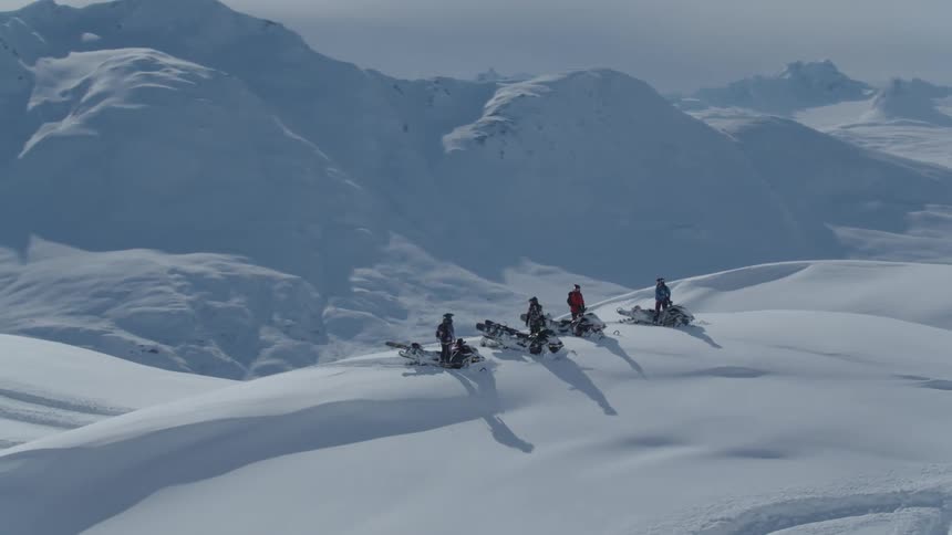 Four people snowmobiling on a hill surrounded by mountains with layers of snow