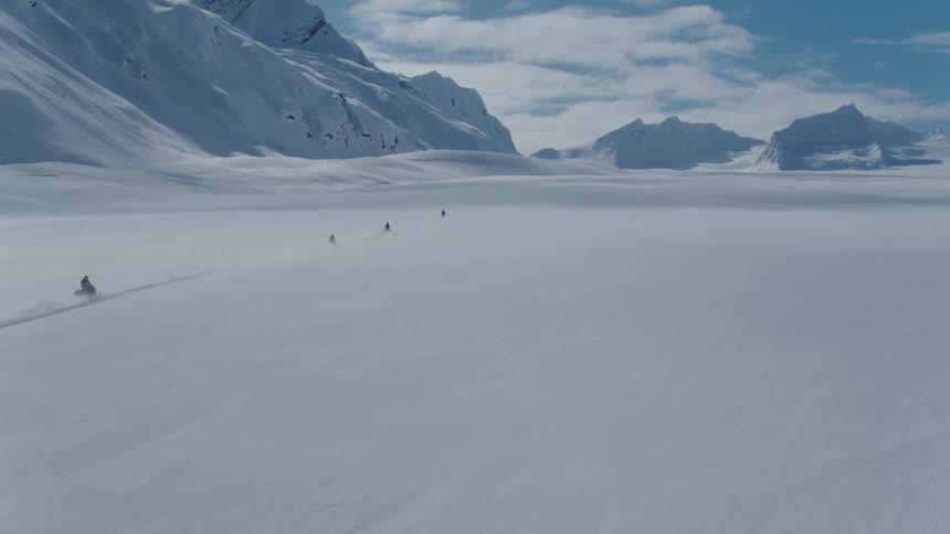 Several people riding snowmobiles in a huge snowcovered area next to large mountains