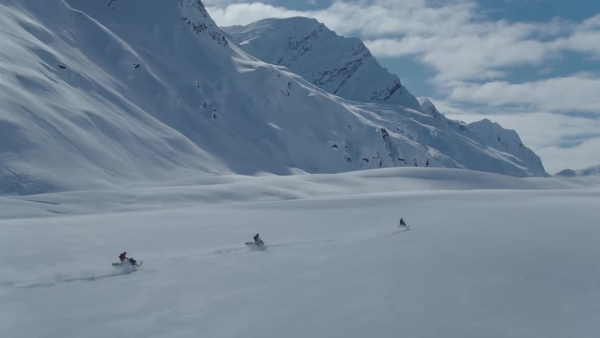 People ride snowmobiles across a snowy plain near snow covered mountains on a cloudy winter day