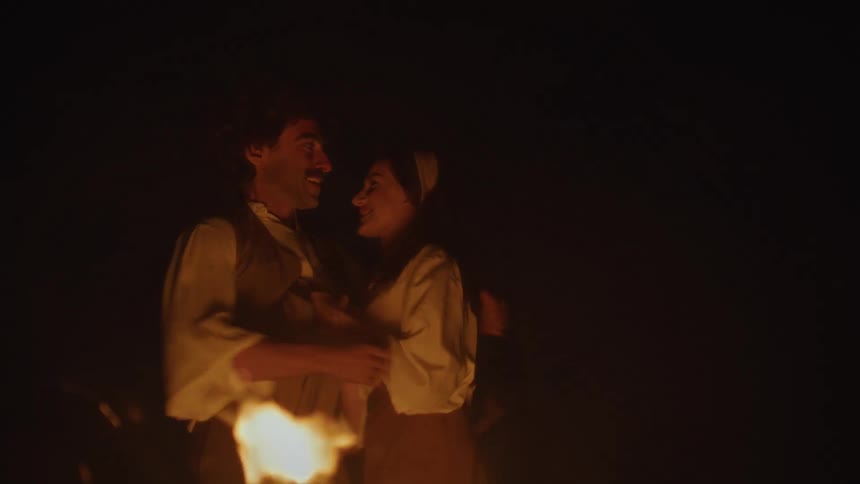 Woman playfully tugs on a scarf before she and a man hug near a campfire at night