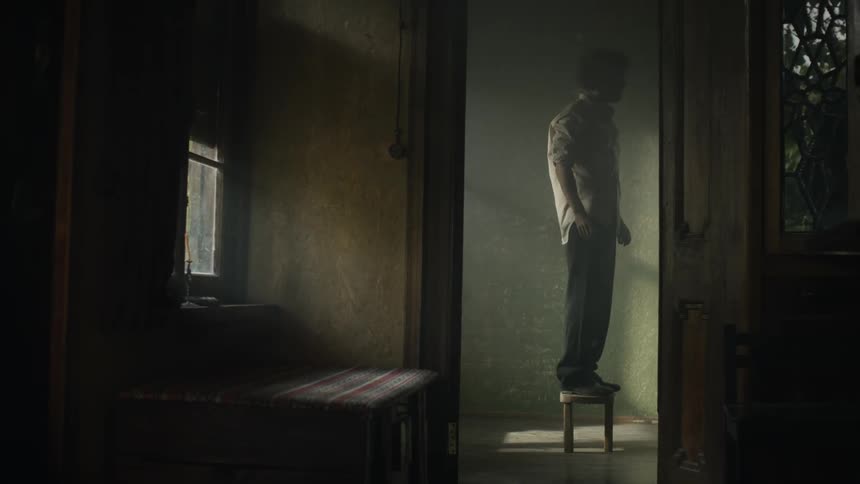Man standing on a step stool in a dimly lit room puts a rope around his neck