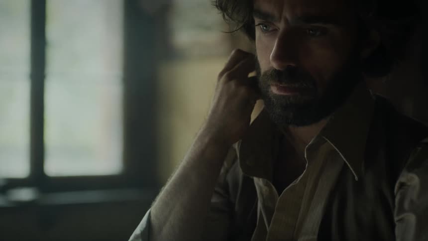 A bearded man is holding his necklace and he is in thoughtful tones as he looks forward