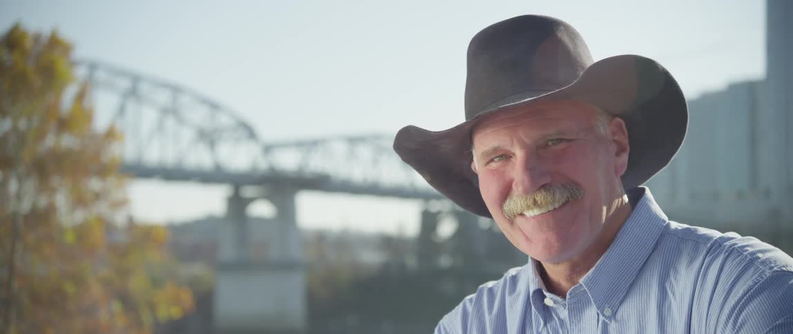 A smiling man wearing a cowboy hat with a bridge and large building in the background