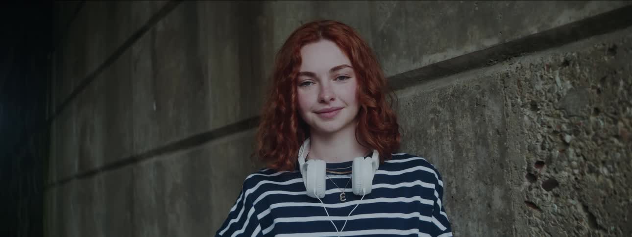 A young woman with red hair and white headphones around her neck smiles while leaning against a concrete wall