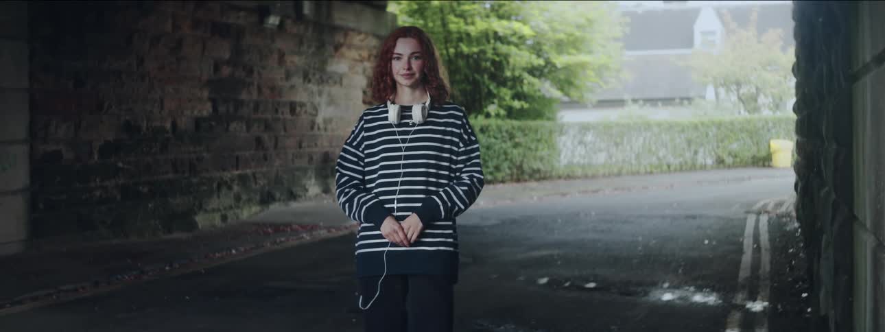 A young woman with red hair and white headphones around her neck smiling while standing in the middle of a road in an underpass