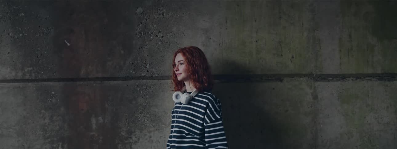 A young woman with red curly hair is standing next to a concrete wall and she has headphones around her neck