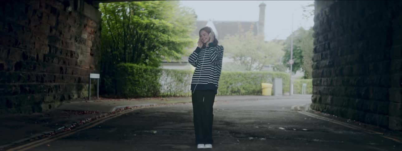 A young woman in a striped shirt has her hands on her headphones and is happy as she listens to music and moves her body