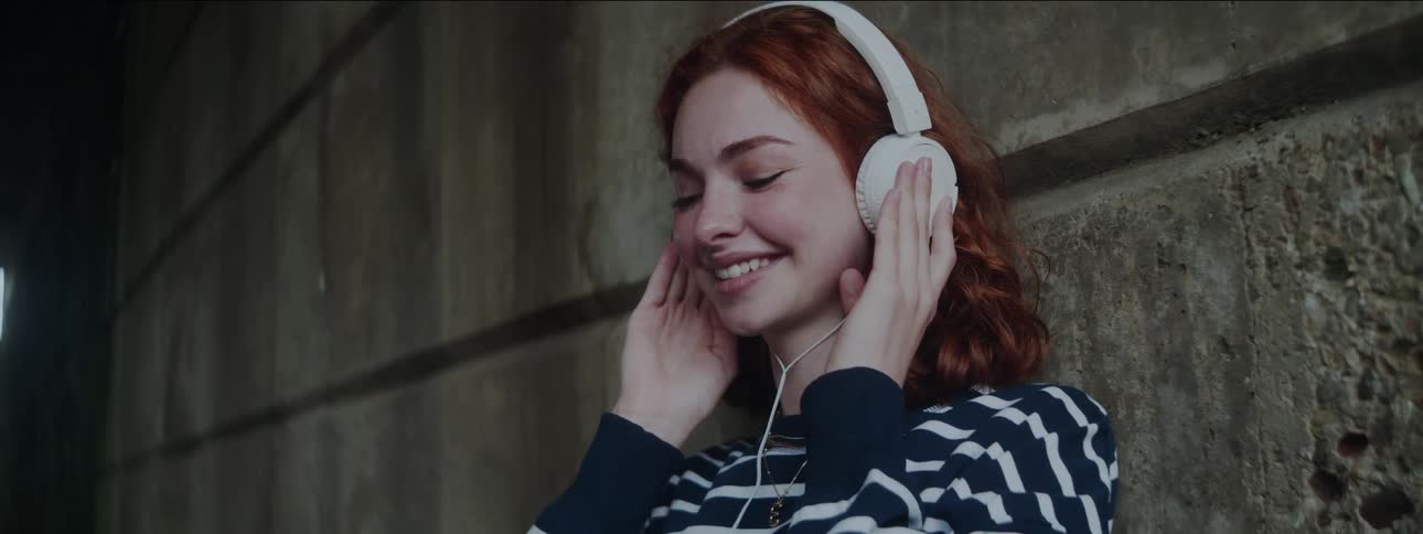A woman with wavy red hair and a striped shirt listens to music through white headphones