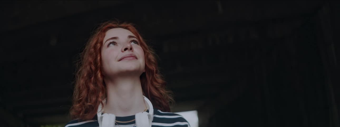 A young woman with red hair and white headphones around her neck smiles while looking up in an underpass