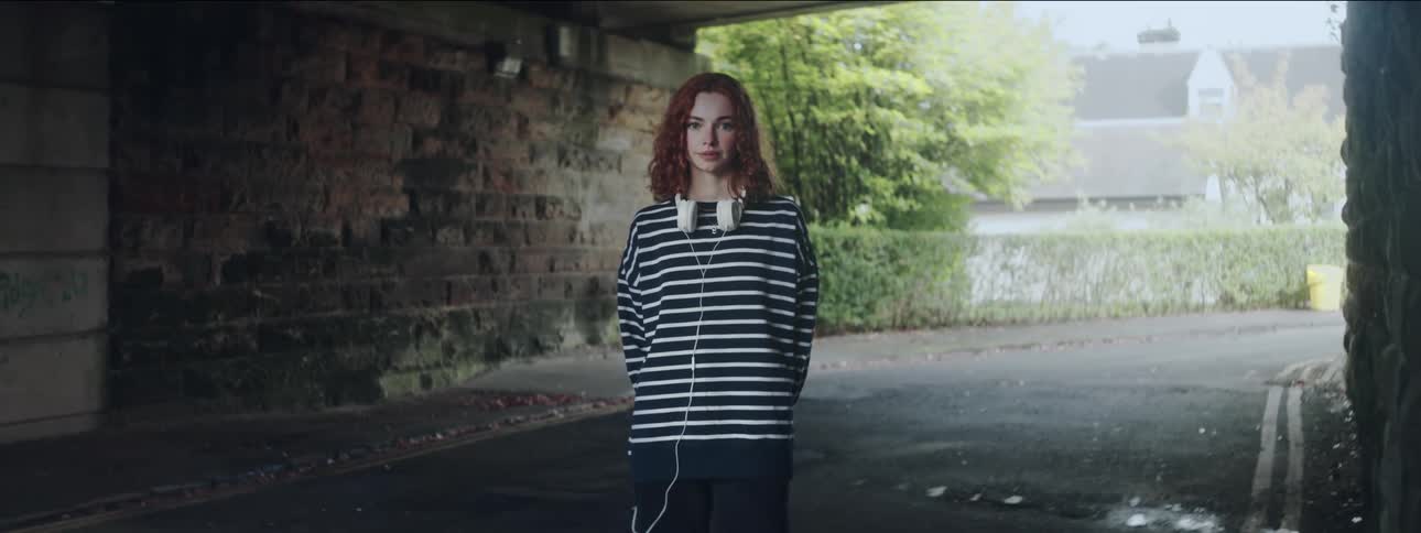 Redhaired woman with curly hair wearing a longsleeved striped tshirt while standing under an overpass with urban houses in the background