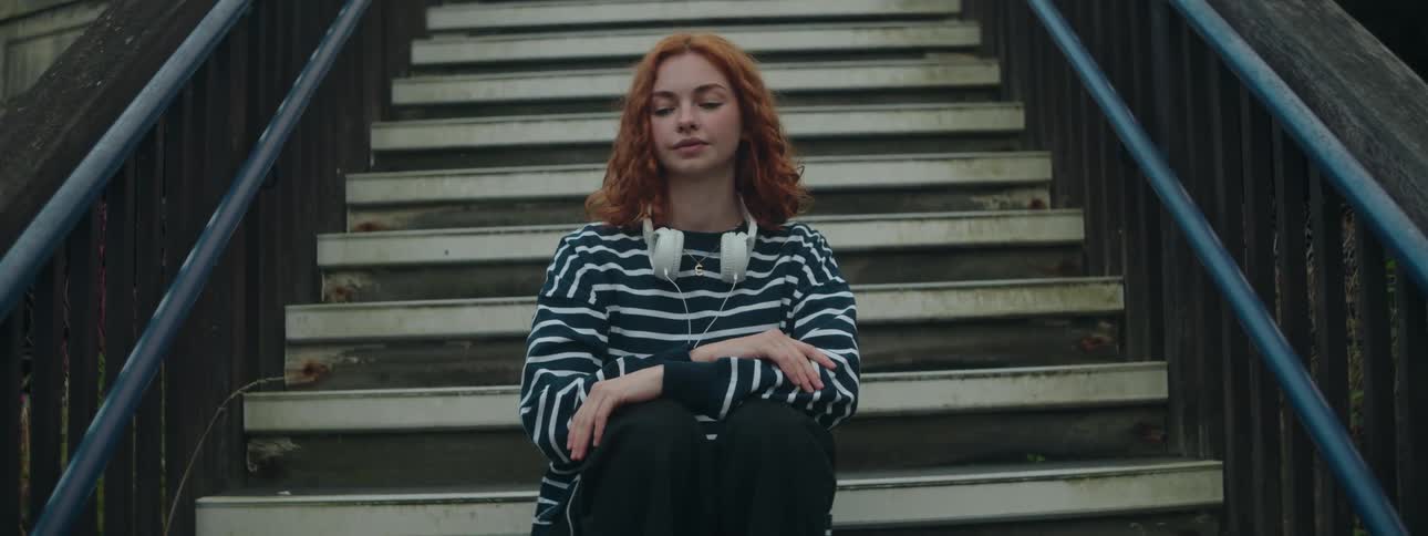 A woman with red hair white headphones and a stripped shirt sits peacefully on stairs eyes closed embodying tranquility and contemplation in a serene moment