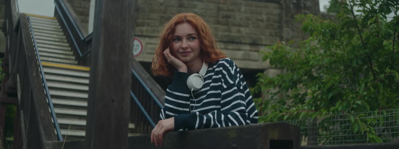 Redhead woman with white headphones and leaning on stairs in an outdoor city setting