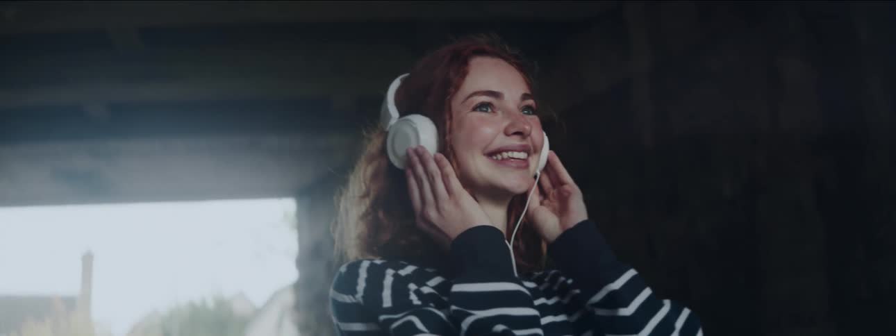 A closeup of a young redhaired teenager listening to music on white headphones under a bridge