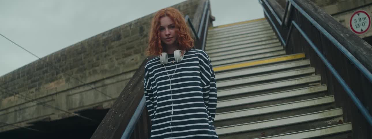A woman with red hair wearing white headphones and a striped shirt stands on the overpass stairs looking down