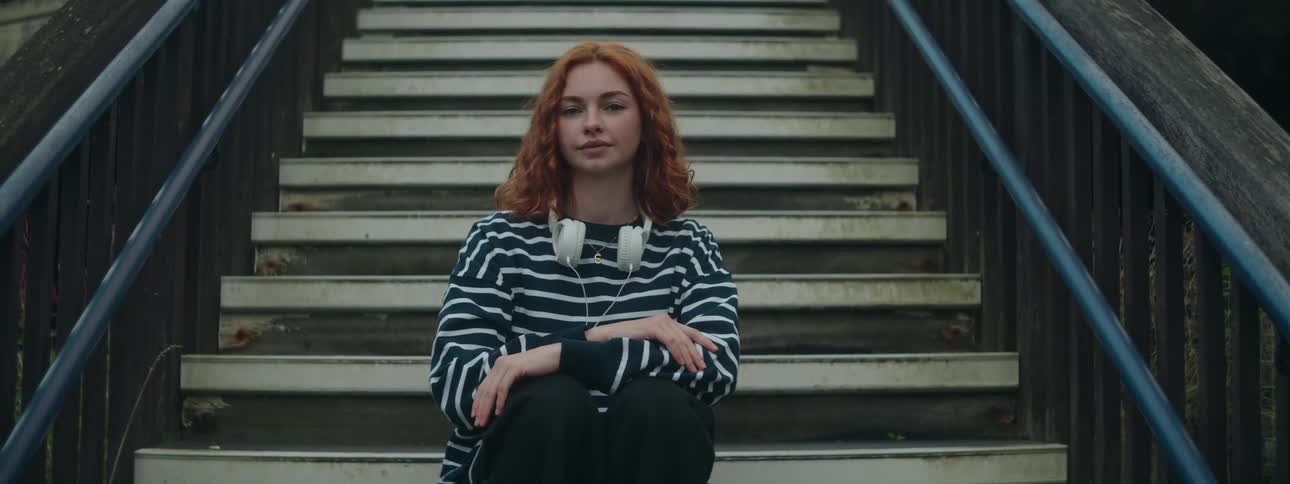 A redheaded young woman sitting in a staircase with white headphones around her neck