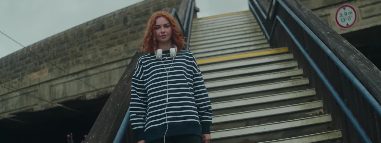 Redhaired woman with white headphones around her neck and wearing a striped longsleeved blouse while standing on the stairs in a public place