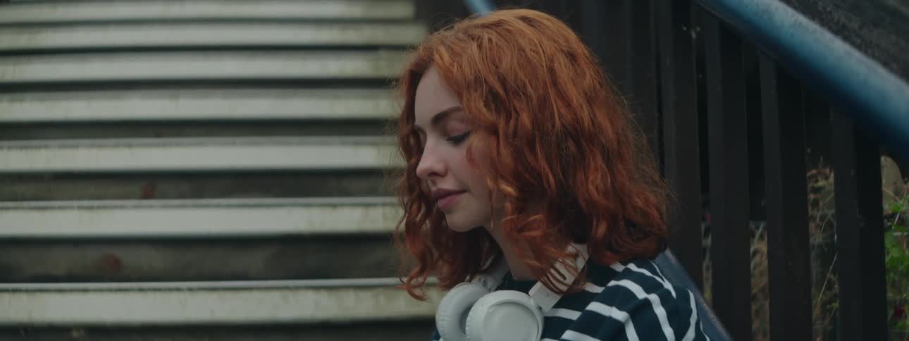 Side view of a curly haired redhead woman with white headphones around her neck sitting on a staircase